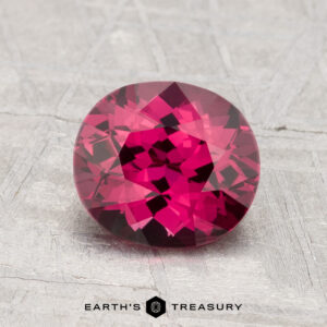 A 1.76-carat round, faceted deep pink spinel is displayed on a textured gray surface with the text "EARTH'S TREASURY" below.