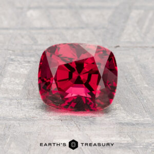 A 1.62-carat cushion-cut red spinel displayed on a textured gray surface, with the text "EARTH'S TREASURY" beneath the gemstone.