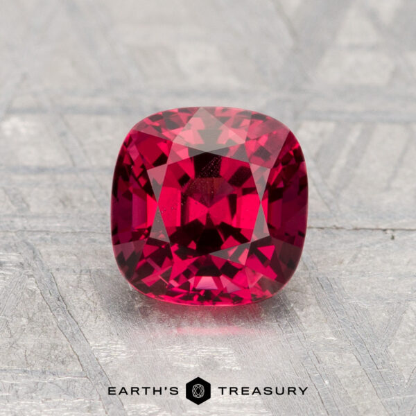 1.33-Carat Spinel A 1.33-carat cushion-cut red spinel rests on a textured gray surface, with the "Earth's Treasury" logo and text at the bottom.