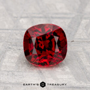 A 1.49-carat cushion-cut spinel rests on a textured gray surface, with "Earth's Treasury" displayed at the bottom.
