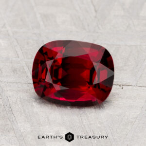 A cushion-cut 1.36-carat Spinel gemstone is displayed on a textured light gray surface, with the text "EARTH'S TREASURY" underneath.