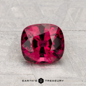 A 1.61-carat cushion-cut red Spinel is displayed on a light gray textured surface with "EARTH'S TREASURY" below.