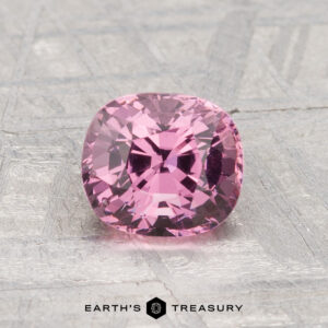 A 1.44-carat cushion-cut pink spinel is displayed on a metallic, textured surface with "EARTH'S TREASURY" at the bottom.