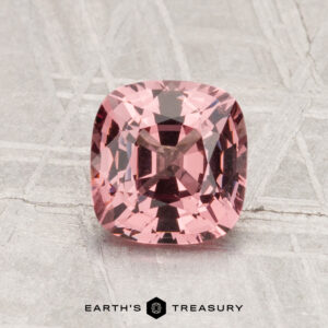 A 1.46-carat, cushion-cut pink spinel is displayed on a light, patterned surface with the text "EARTH'S TREASURY" below it.