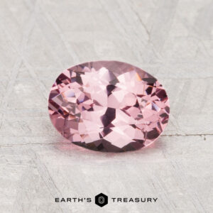 A 1.36-carat faceted oval pink spinel is displayed on a light gray textured surface with the text "EARTH'S TREASURY" below it.