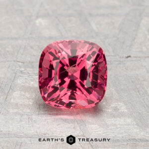 A 1.26-carat cushion-cut reddish-pink spinel is displayed on a textured gray surface, with "Earth's Treasury" visible at the bottom.