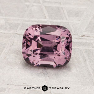 A 1.49-carat cushion-cut purple Spinel is displayed on a textured, light gray surface with the text "EARTH'S TREASURY" below.