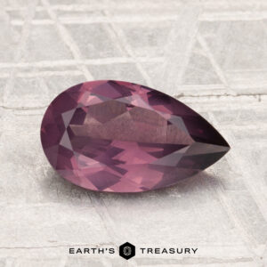 A faceted, pear-shaped 1.56-carat spinel gemstone is displayed on a textured light-gray surface with "Earth's Treasury" branding at the bottom.