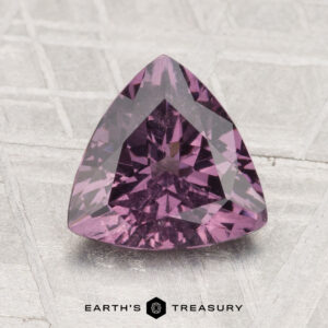 A 1.87-carat purple spinel, cut in a triangular shape, rests on a light textured surface with the text "EARTH'S TREASURY" beneath it.