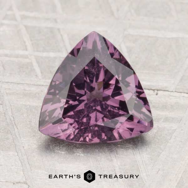 1.87-Carat Spinel A 1.87-carat purple spinel, cut in a triangular shape, rests on a light textured surface with the text "EARTH'S TREASURY" beneath it.
