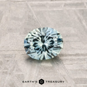 A 0.79-carat Montana Sapphire, round and faceted with pale blue-green hues, rests on a textured gray surface. "EARTH'S TREASURY" is displayed at the bottom of the image.