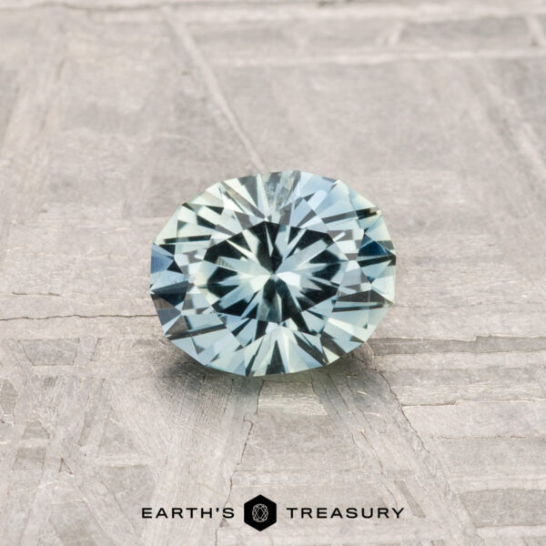 0.79-Carat Montana Sapphire A 0.79-carat Montana Sapphire, round and faceted with pale blue-green hues, rests on a textured gray surface. "EARTH'S TREASURY" is displayed at the bottom of the image.