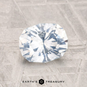 A 1.78-carat, round brilliant-cut Montana Sapphire is displayed on a textured gray surface, with the text "EARTH'S TREASURY" beneath it.