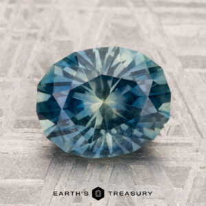 A 2.90-carat round, faceted Montana sapphire in blue-green is displayed on a textured metallic surface with "EARTH'S TREASURY" at the bottom.