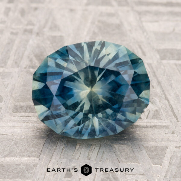 2.90-Carat Montana Sapphire A 2.90-carat round, faceted Montana sapphire in blue-green is displayed on a textured metallic surface with "EARTH'S TREASURY" at the bottom.