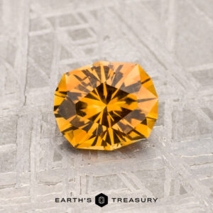 A 1.02-carat faceted round Montana sapphire with an orange hue is displayed on a textured gray surface, with the text "Earth's Treasury" below.