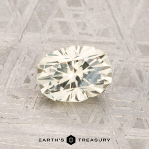 A 1.14-carat faceted oval pale yellow Montana sapphire is displayed on a textured gray surface with the text "EARTH'S TREASURY" at the bottom.