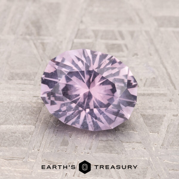 1.53-Carat Lilac Pink Montana Sapphire The 1.53-Carat Lilac Pink Montana Sapphire, oval-cut and faceted, is displayed on a textured light gray surface with the Earth’s Treasury logo below.