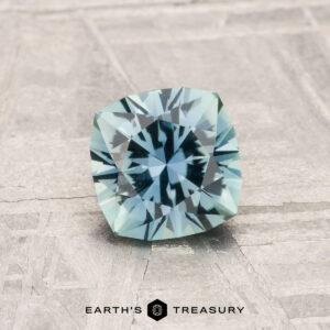 A 1.05-Carat Montana Sapphire in cushion cut and light aqua blue is shown on a textured surface with "EARTH'S TREASURY" text at the bottom.