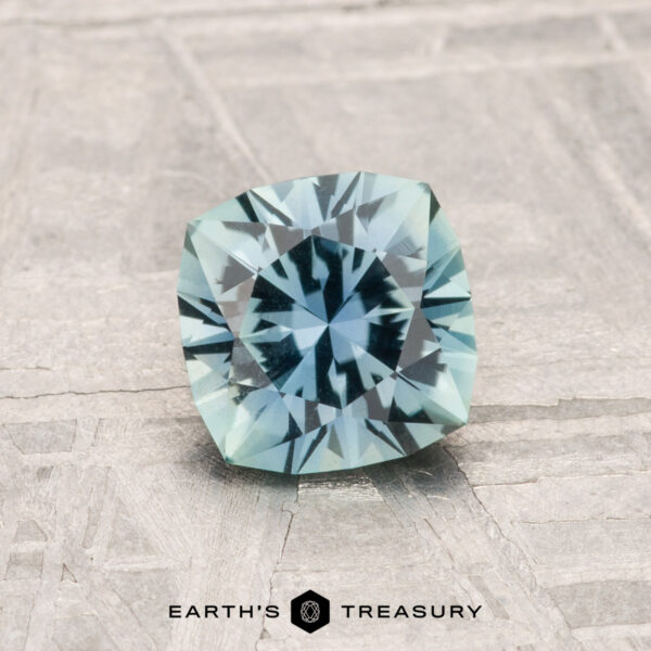 1.05-Carat Montana Sapphire A 1.05-Carat Montana Sapphire in cushion cut and light aqua blue is shown on a textured surface with "EARTH'S TREASURY" text at the bottom.