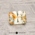 A 1.14-carat Montana sapphire, cut in a rectangular faceted shape with yellow and orange tones, rests on a textured gray surface; "EARTH'S TREASURY" is displayed below.