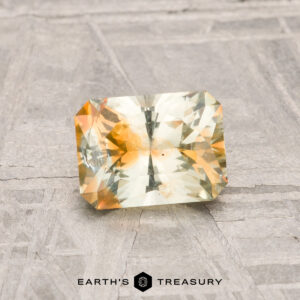 A 1.14-carat Montana sapphire, cut in a rectangular faceted shape with yellow and orange tones, rests on a textured gray surface; "EARTH'S TREASURY" is displayed below.