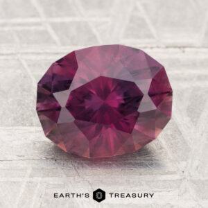 A 3.06-carat round, faceted Montana sapphire in a purple-red hue is displayed on a textured surface with the text "EARTH'S TREASURY" below it.