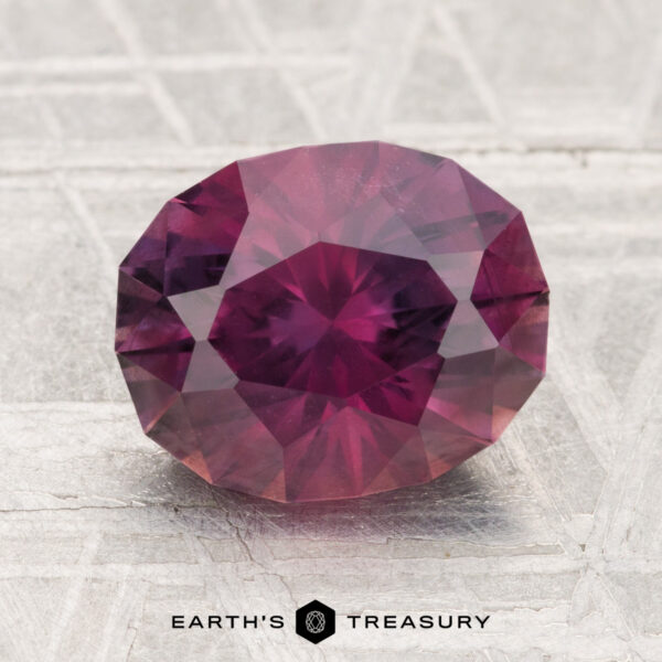 3.06-Carat Montana Sapphire A 3.06-carat round, faceted Montana sapphire in a purple-red hue is displayed on a textured surface with the text "EARTH'S TREASURY" below it.