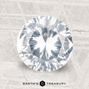 A round, faceted 2.77-carat sapphire displayed on a textured light surface with "EARTH'S TREASURY" at the bottom.