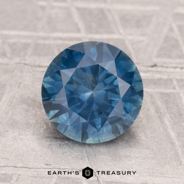 2.05-Carat Montana Sapphire (Heated) A round, faceted 2.05-carat heated Montana sapphire is shown on a textured gray surface with "EARTH'S TREASURY" beneath it.