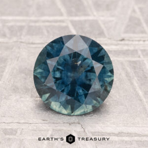 1.84-carat heated Montana sapphire, round brilliant-cut in blue-green, displayed on a textured gray surface with "EARTH'S TREASURY" text at the bottom.
