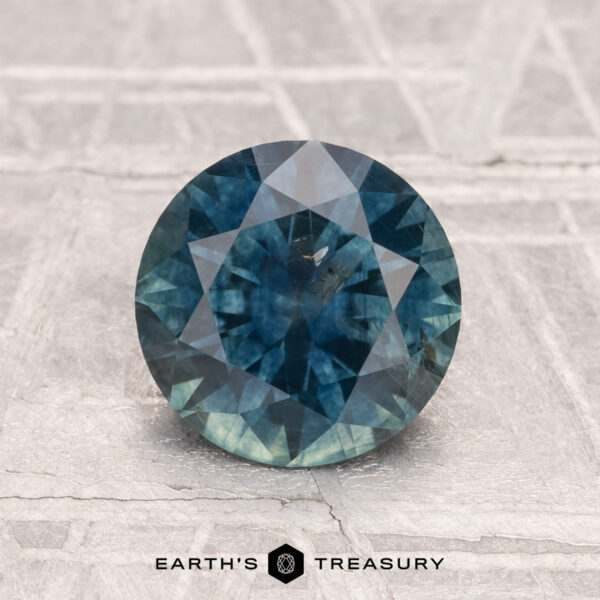 1.84-Carat Montana Sapphire (Heated) 1.84-carat heated Montana sapphire, round brilliant-cut in blue-green, displayed on a textured gray surface with "EARTH'S TREASURY" text at the bottom.