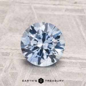 A 1.60-carat heated Montana sapphire with a light blue round brilliant cut is displayed on a textured, light background. The Earth's Treasury logo is visible at the bottom of the image.