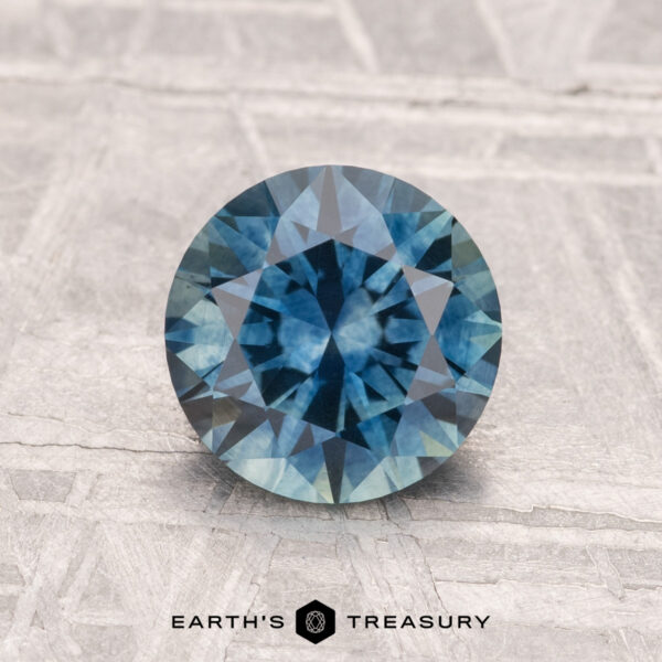 1.59-Carat Montana Sapphire (Heated) A 1.59-carat heated Montana Sapphire with a round brilliant cut sparkles on a textured light gray background, with "EARTH'S TREASURY" displayed at the bottom.