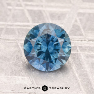 A vivid blue 1.59-carat heated Montana Sapphire, round brilliant-cut, sits on a textured light surface with the "Earth’s Treasury" logo and text shown at the bottom.