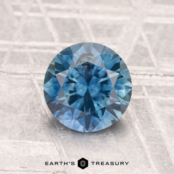 1.59-Carat Montana Sapphire (Heated) A vivid blue 1.59-carat heated Montana Sapphire, round brilliant-cut, sits on a textured light surface with the "Earth’s Treasury" logo and text shown at the bottom.