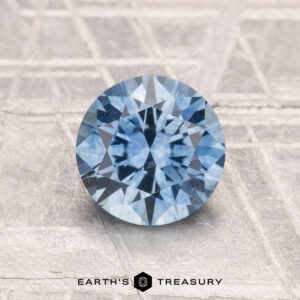1.50-Carat Montana Sapphire (Heated) in round brilliant cut displayed on a textured light gray surface, with "Earth's Treasury" logo and text at the bottom.