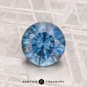 A round, faceted 1.48-carat heated Montana sapphire is displayed on a light textured surface with the text “EARTH’S TREASURY” beneath it.
