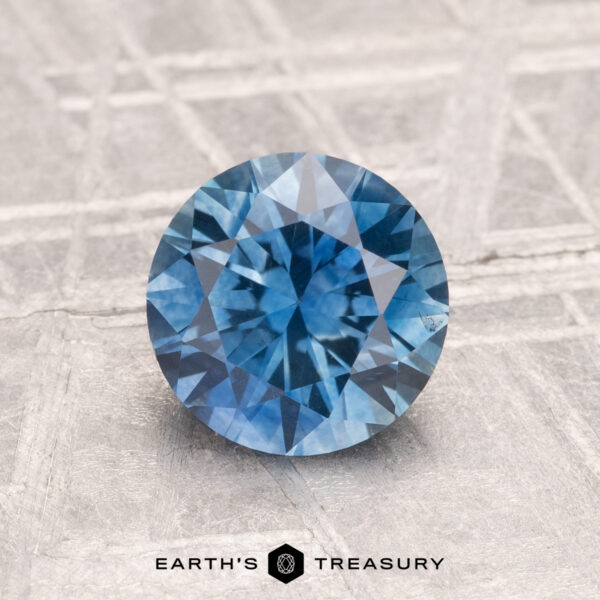 1.46-Carat Montana Sapphire (Heated) A round, faceted 1.46-carat heated Montana sapphire in blue shown on a textured gray background with "EARTH'S TREASURY" written at the bottom.