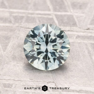 A 1.42-carat Montana Sapphire (Heated), round and faceted with a pale green hue, is shown on a textured light gray surface, featuring the text "Earth's Treasury" at the bottom.
