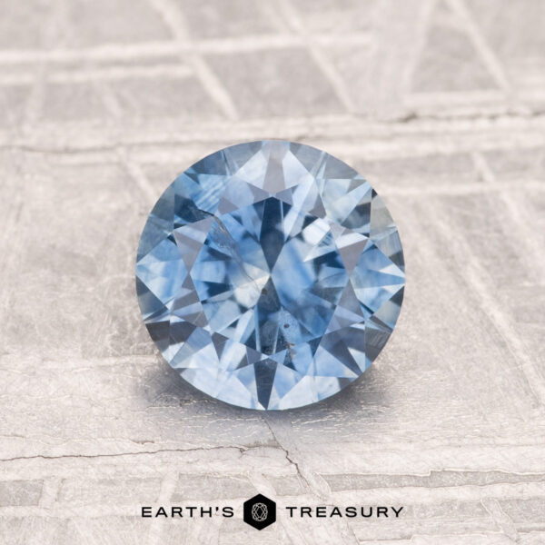 1.37-Carat Montana Sapphire (Heated) A 1.37-carat round brilliant-cut heated Montana sapphire sits on a textured light gray surface, with "EARTH'S TREASURY" displayed at the bottom.