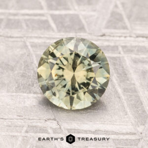 A round, faceted 1.35-carat heated Montana sapphire with a greenish-yellow hue is displayed on a textured gray surface featuring the "Earth's Treasury" logo and text at the bottom.