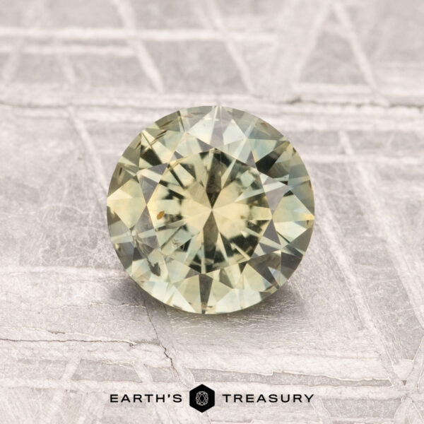 1.35-Carat Montana Sapphire (Heated) A round, faceted 1.35-carat heated Montana sapphire with a greenish-yellow hue is displayed on a textured gray surface featuring the "Earth's Treasury" logo and text at the bottom.