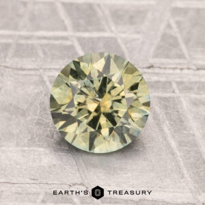 A 1.35-carat heated Montana sapphire, round and faceted with yellow-green hues, is displayed on a textured surface with "EARTH'S TREASURY" at the bottom.