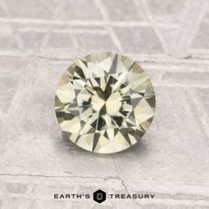 A 1.30-carat heated Montana sapphire, round and faceted in pale yellow, is shown on a textured gray surface with "Earth's Treasury" beneath it.