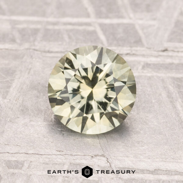 1.30-Carat Montana Sapphire (Heated) A 1.30-carat heated Montana sapphire, round and faceted in pale yellow, is shown on a textured gray surface with "Earth's Treasury" beneath it.