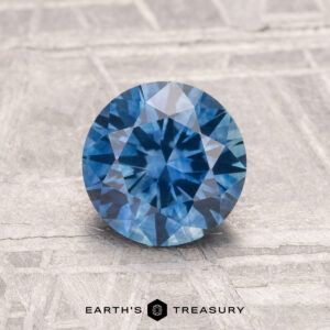 A 1.30-carat heated Montana Sapphire, round brilliant-cut and blue in color, is displayed on a textured light gray surface with "EARTH'S TREASURY" at the bottom.