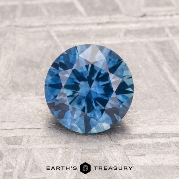 1.30-Carat Montana Sapphire (Heated) A 1.30-carat heated Montana Sapphire, round brilliant-cut and blue in color, is displayed on a textured light gray surface with "EARTH'S TREASURY" at the bottom.