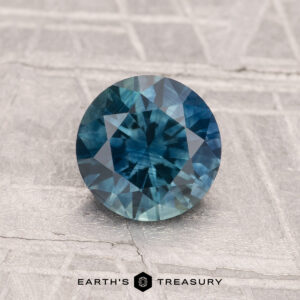 A 1.26-carat heated Montana sapphire, round faceted and blue-green in color, rests on a textured light gray surface with the "Earth's Treasury" logo and text displayed at the bottom.