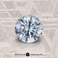 A 1.26-carat heated Montana sapphire, cut in a round brilliant shape, rests on a textured gray surface with "Earth's Treasury" displayed below.